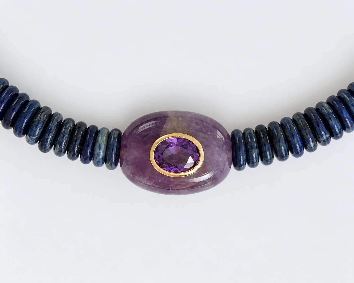 A beaded necklace featuring a central oval purple amethyst bead with a gold bezel setting, flanked by dark blue lapis lazuli beads on either side, shown against a plain white background.