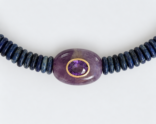 A beaded necklace featuring a central oval purple amethyst bead with a gold bezel setting, flanked by dark blue lapis lazuli beads on either side, shown against a plain white background.