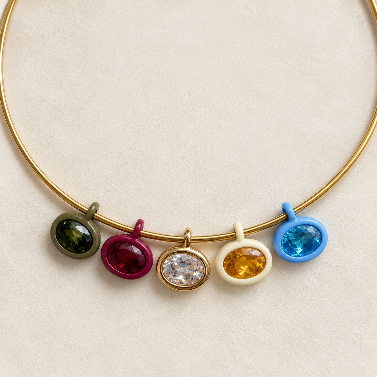 A gold-tone circular bangle necklace with five dangling oval gemstone charms in olive green, magenta, clear, yellow, and blue, displayed on a textured cream background.