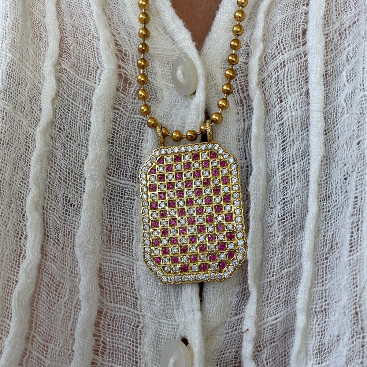 A close-up of a gold beaded necklace with a rectangular pendant covered in red and white stones, worn over a white textured blouse.