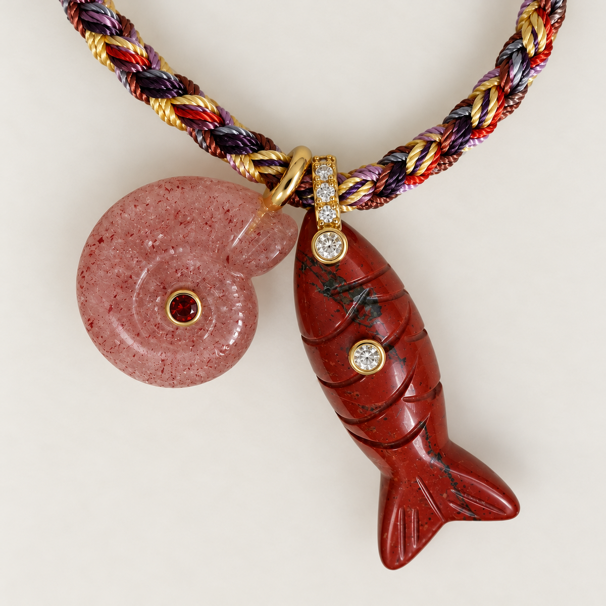 A close-up of a jewelry piece featuring a carved red fish-shaped charm with small clear stones, a round pink stone with a red center, and a braided cord in red, gold, purple, and white threads, set against a plain light background.