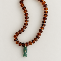 A beaded necklace made of translucent amber-brown round beads with a small green fish-shaped pendant and gold-tone clasp, photographed on a plain light background.