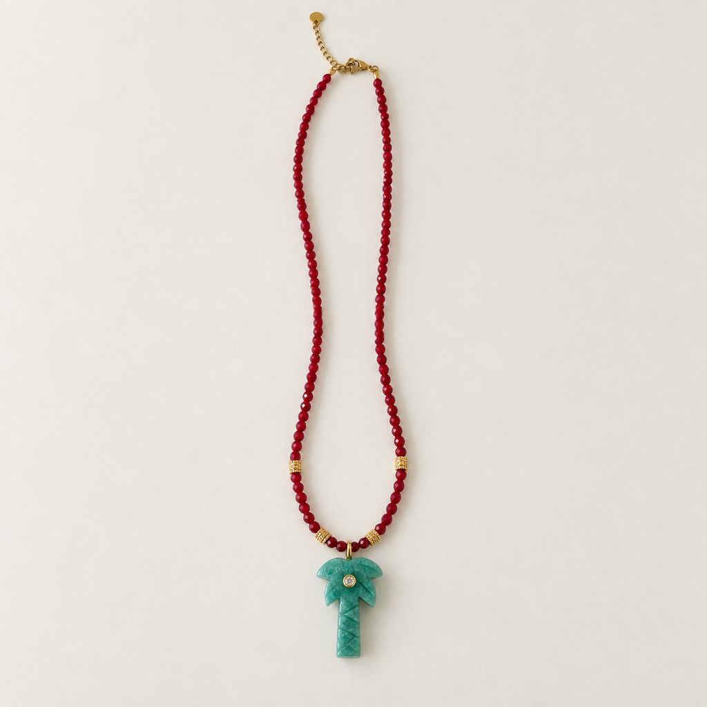 A beaded necklace featuring a long strand of red faceted beads, small gold spacer beads, and a carved turquoise palm tree pendant with a central white pearl accent, displayed on a clean off-white background.