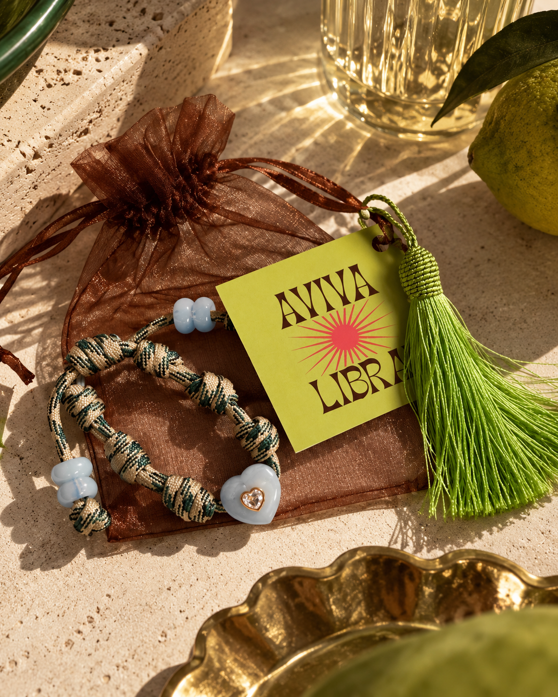 A braided bracelet with light blue beads and a light blue heart charm rests on a brown sheer drawstring pouch. A bright yellow tag reading 'AVVA LIBRA' with a sunburst logo is attached, along with a green tassel.