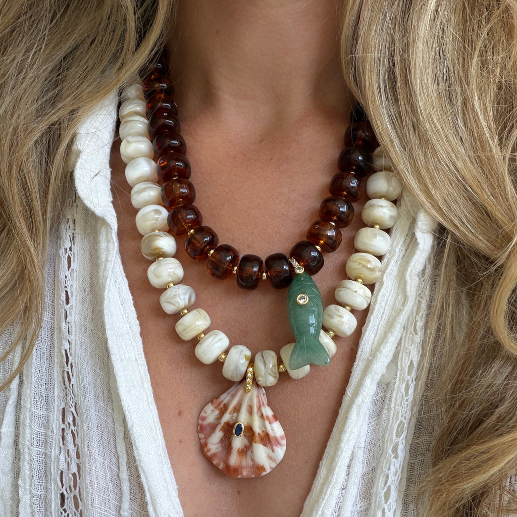 A close-up of a woman wearing a bohemian-style layered necklace. The necklace features dark amber brown beads, cream-colored stone beads, a green jade fish charm, a pink and white seashell pendant, and small gold spacer rings, all worn over a white lace-trim blouse.