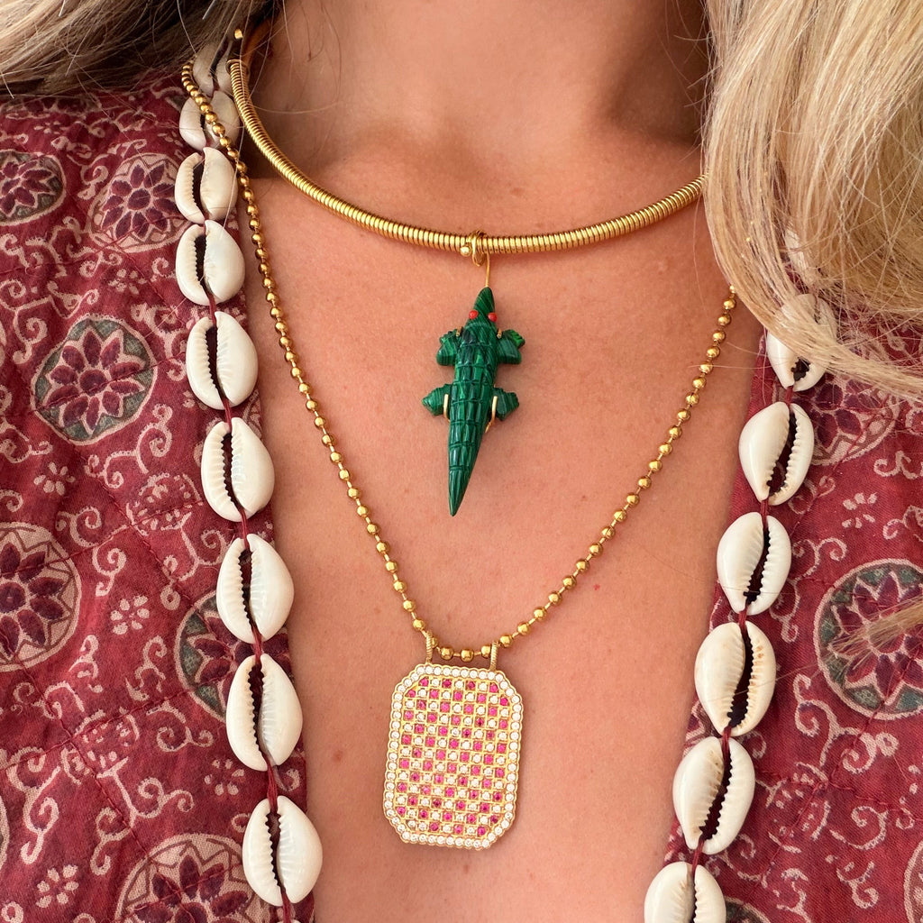 A close-up of a model wearing layered necklaces. The center necklace features a green crocodile pendant on a gold chain, layered over a long gold chain with a rectangular gold and white patterned pendant.