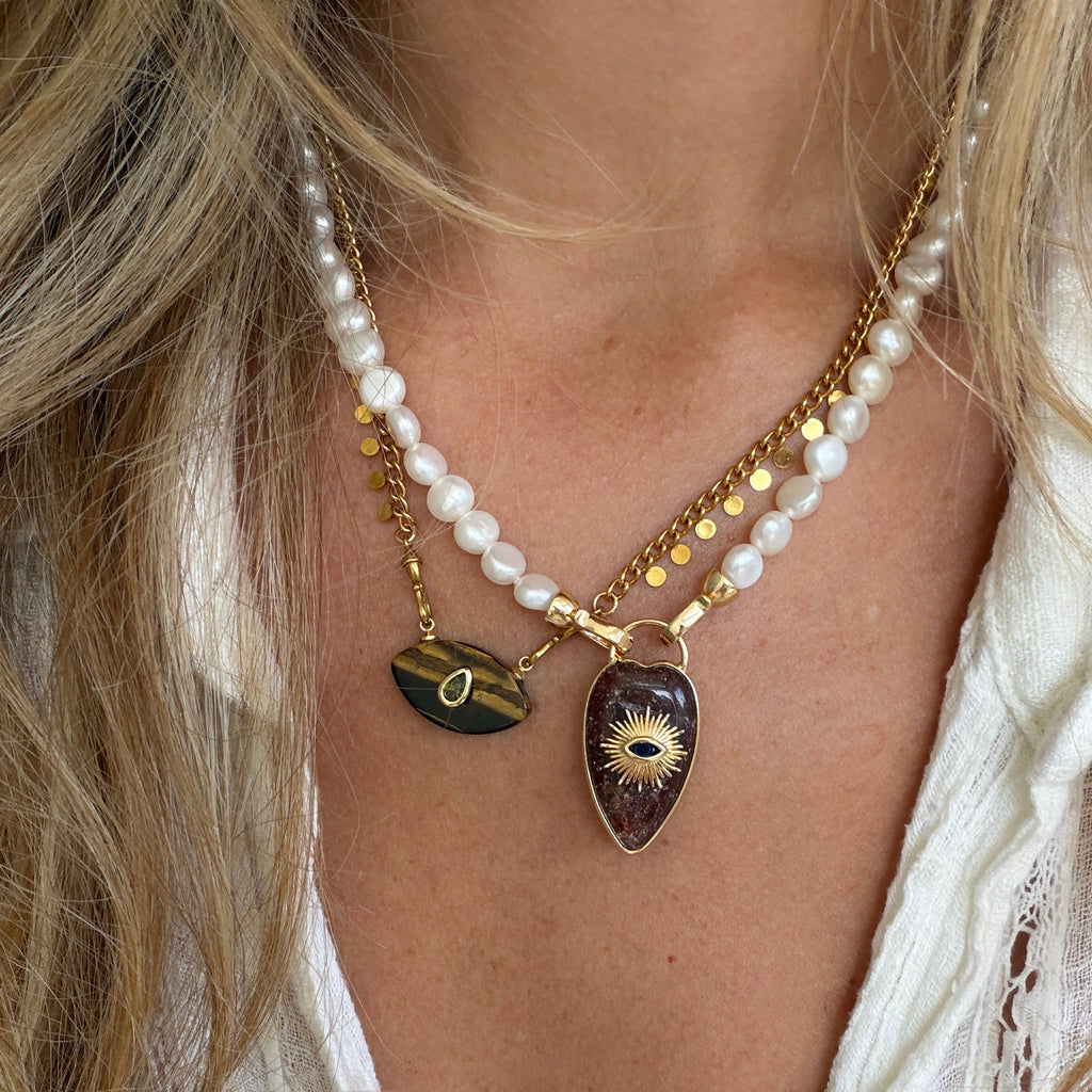 A close-up of a woman wearing a layered necklace with white pearl beads, gold chains, and two teardrop-shaped pendants: one dark green tiger's eye and one dark brown wood with a gold eye design.