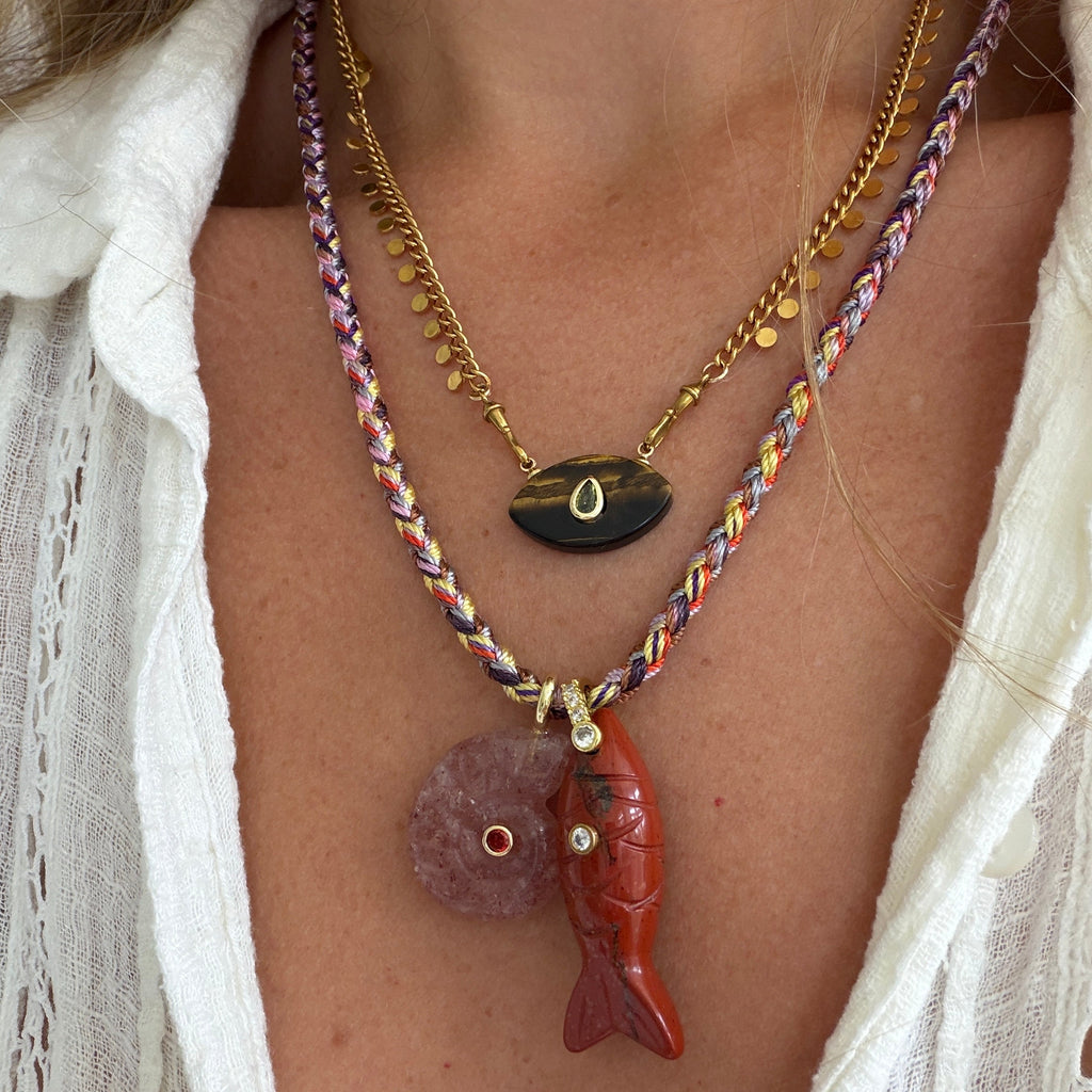 A close-up of a person wearing a layered necklace set. The set includes a gold chain with a black onyx pendant, a multicolor braided cord necklace with a red carved fish pendant and a pink stone pendant, and a thin gold chain with small gold beads.