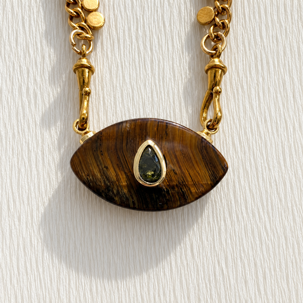 A close-up of a gold-tone necklace with an oval brown tiger's eye pendant featuring a small teardrop-shaped green gemstone in the center, set against a textured white background.