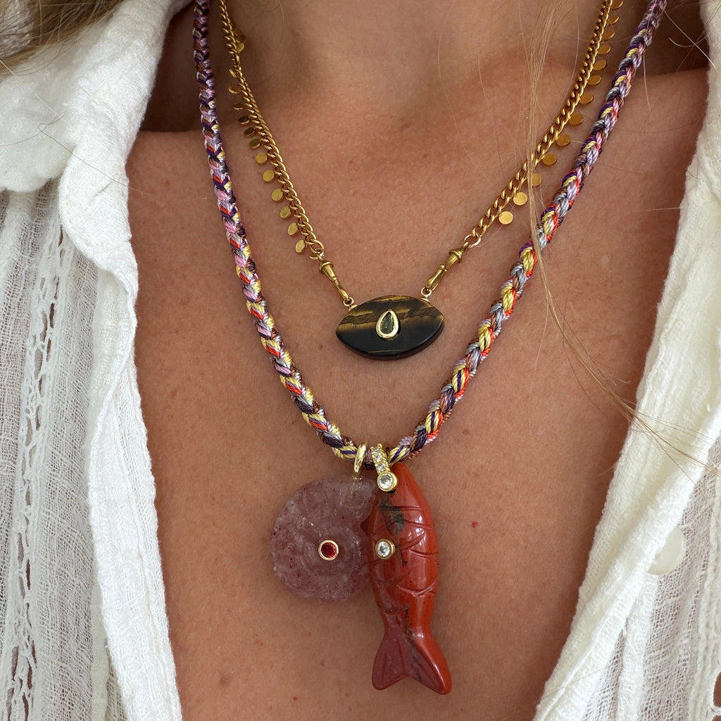 A close-up of a person wearing a layered necklace set. The set includes a gold chain with a black onyx pendant, a multicolor braided cord necklace with a red carved fish pendant and a pink stone disc, and a thin gold chain with small gold beads.