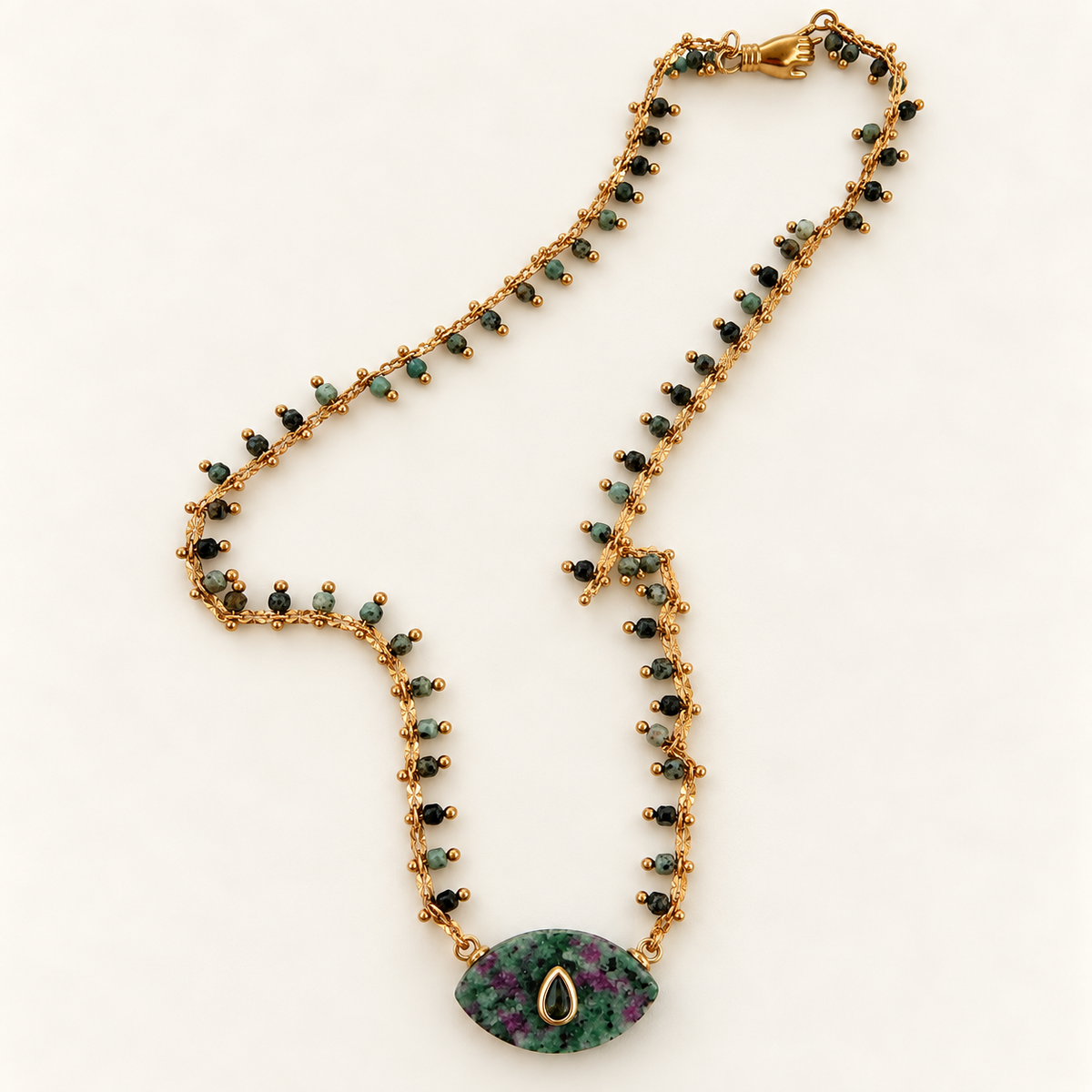 A gold-tone beaded necklace with small green and black beads, featuring a teardrop-shaped pendant made from green and purple mottled stone with a gold teardrop setting, displayed on a clean white background.