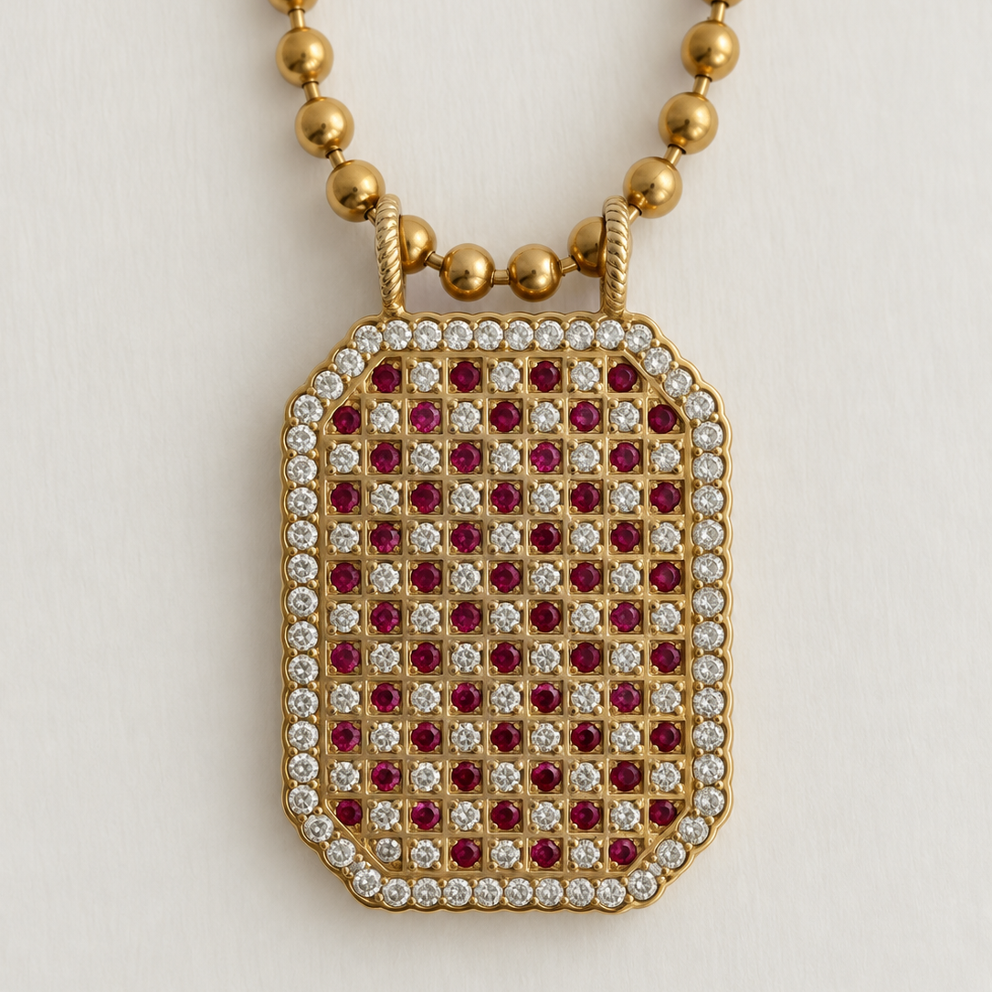 A gold pendant necklace featuring a rectangular dog-tag style pendant encrusted with red and clear stones, suspended from a gold beaded chain against a light background.