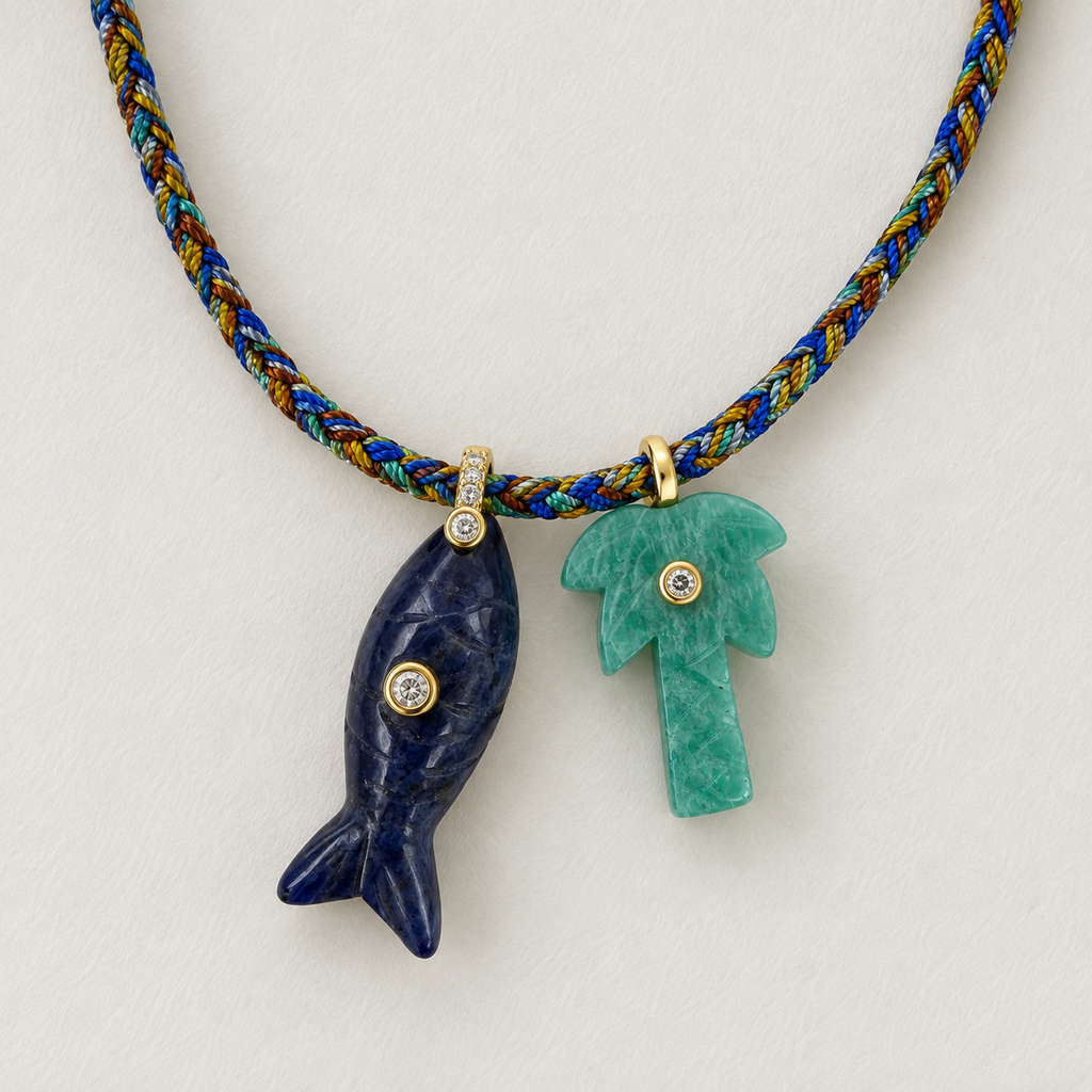 A close-up of a necklace with a braided cord in blue, gold, and green, featuring two carved stone pendants: a dark blue fish-shaped pendant and a green palm tree pendant, both with small gold and clear stone accents, on a light off-white background.