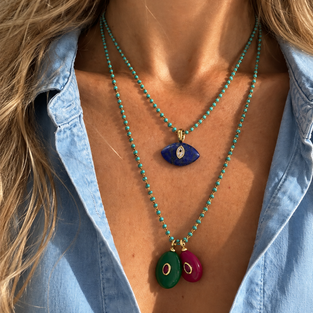 A close-up of a model wearing two layered turquoise beaded necklaces. The top necklace features a blue evil eye pendant, and the longer bottom necklace features two oval pendants, one green and one magenta pink, both with gold-tone borders and white centers.