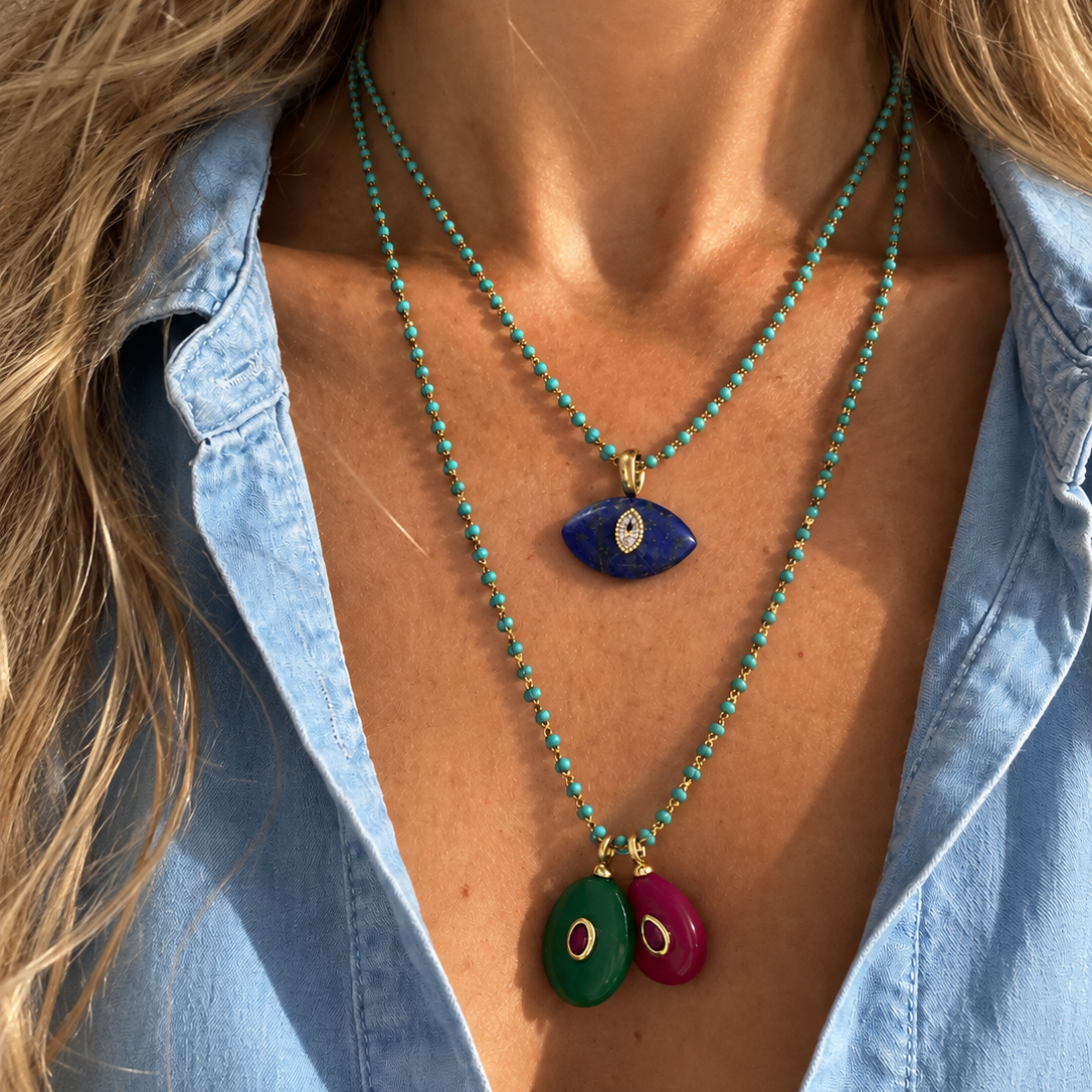A close-up of a model wearing two layered turquoise beaded necklaces. The top necklace features a blue evil eye pendant, and the longer bottom necklace features two oval pendants, one green and one magenta pink, both with gold-tone borders and white centers.