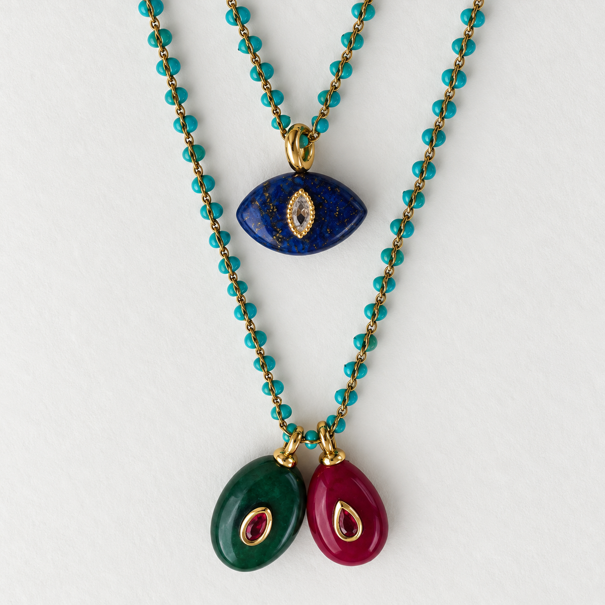 A close-up of a multi-colored beaded necklace with three distinct drop chains: one in vibrant turquoise, one in deep blue with gold flecks and an evil eye motif, and one in dark green with an evil eye motif. The top of the necklace features two large teardrop-shaped beads, one magenta and one dark green, both accented with gold and a small red gemstone.
